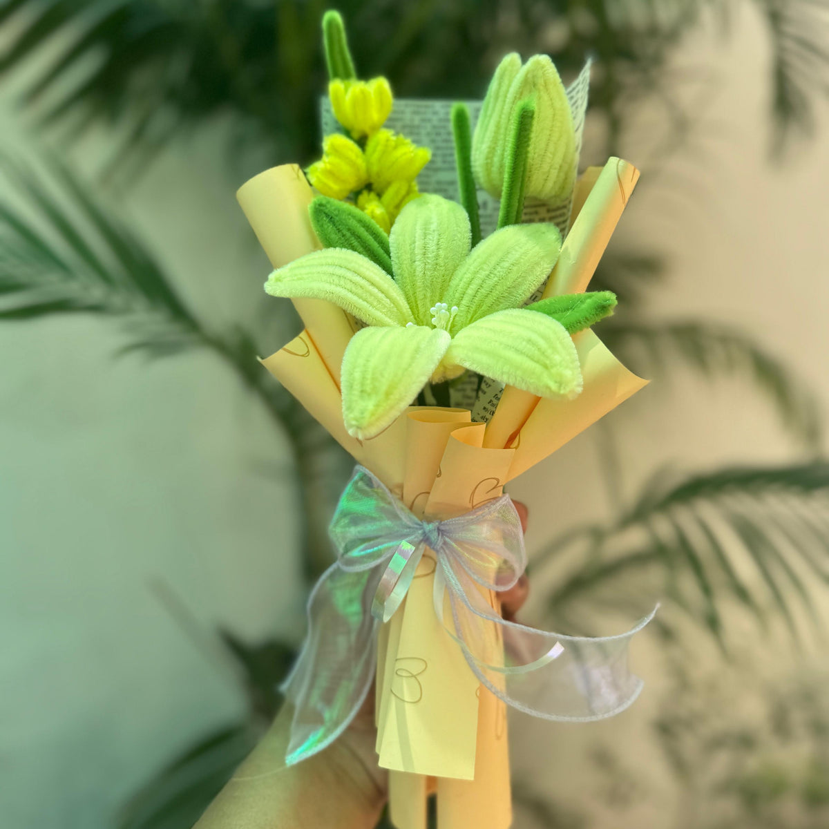Bouquet of artificial flowers wrapped in yellow paper with a green bow, held against a blurred green plant background.